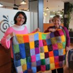 Concha y Marisol muestran una manta que las tejedoras han unido a partir de «cuadritos» de 10x10cm